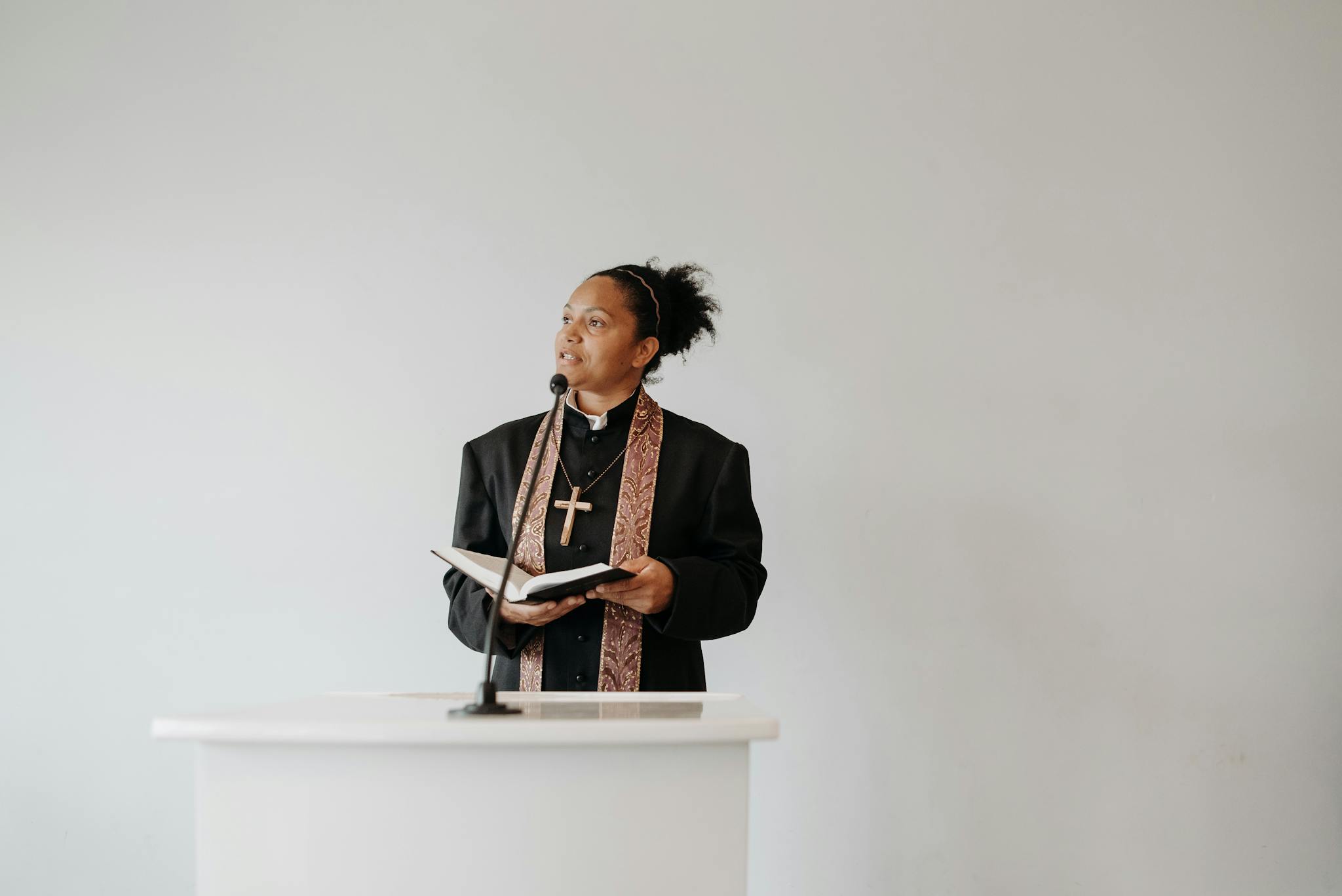 A female pastor delivers a sermon at a church podium, holding a Bible and wearing a cross.
